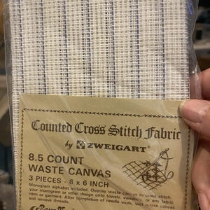 May include: A package of three 6 x 6 inch pieces of 8.5 count Zweigart waste canvas for counted cross stitch. The white fabric features thin vertical blue guide lines. The packaging includes text detailing the product name and usage instructions.