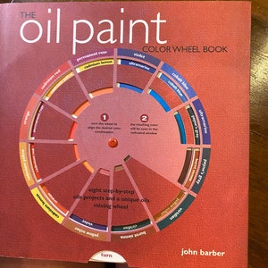 Pode incluir: Um livro intitulado "The Oil Paint Color Wheel Book" com um diagrama de roda de cores. A capa é marrom avermelhado com o título em branco. A roda de cores mostra várias cores de tinta como vermelho cádmio, ultramarino e ocre amarelo.