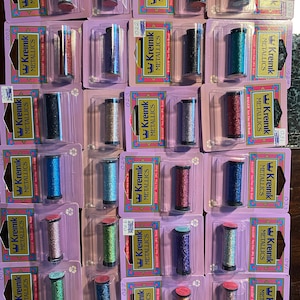 May include: A collection of 20 spools of metallic embroidery floss, each in a different color, arranged in four rows of five spools. The spools are labeled with the brand name "Kreinik" and the number "027".