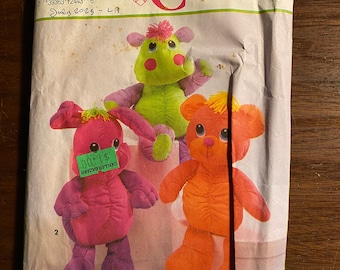 Puede incluir: Sobre de patrón Simplicity Crafts con tres diseños de animales de peluche: una jirafa verde, un oso naranja y un conejo rosa. El sobre tiene el texto "7808 ONE SIZE" y "Crafts". Una etiqueta de precio es visible en el conejo.