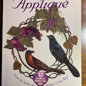 May include: Book cover of "Fancy Appliqué" by Elly Sienkiewicz. It shows a wreath with purple grapes, leaves, and two birds. Text includes "12 Lessons to Enhance Your Skills" and "Appliqué 12 Easy Ways!"