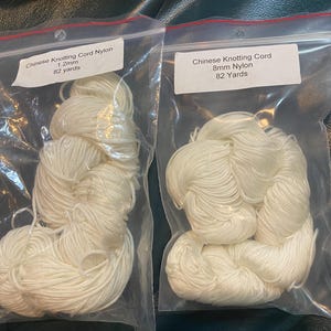May include: Two clear plastic bags containing white Chinese knotting cord. The bags have labels that read "Chinese Knotting Cord Nylon" with measurements of 1.2mm and .8mm, and 82 yards. The cord is tightly wound.