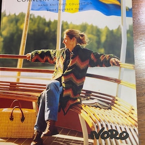 May include: The cover of a knitting book, "CORNELIA TUTTLE HAMILTON HAND KNITTING COLLECTION BOOK NUMBER TWO", shows a woman in a colourful knitted coat. A yellow and blue flag is in the background. A woven basket and the word "NORO" are also visible.