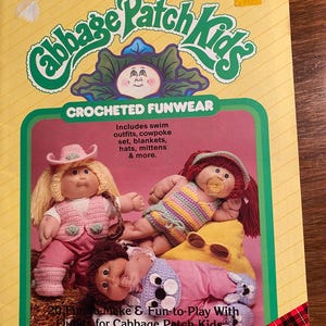 May include: A crochet pattern booklet for Cabbage Patch Kids featuring a red and black plaid design on the cover. The booklet includes instructions for making a variety of crocheted outfits, including swimsuits, cowpoke sets, blankets, hats, and mittens.