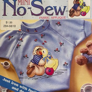 May include: A blue fabric applique featuring a teddy bear and a duckling. The applique is on a light blue shirt with a white collar. The text "No Sew" is printed in red and white on the packaging. The text "Fabric Applique" is printed in black on the packaging. The text "Just iron with fusible web and decorate!" is printed in black on the packaging.