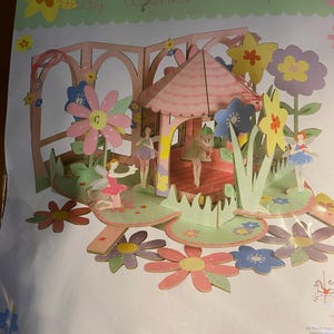 May include: A pink and green papercraft fairy centerpiece with a small pink gazebo and flowers. The fairies are magnetic and movable. The text on the packaging reads "Fairy Wishes Centerpiece" and "Movable Magnetic Fairies. Simple to assemble, no gluing or cutting required."