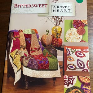 Può includere: Una copertina di rivista con il titolo "BITTERSWEET ART TO HEART" presenta una colorata trapunta drappeggiata su una poltrona in pelle marrone. La trapunta ha un design patchwork con vari motivi e colori. Sono visibili anche zucche decorative e un vaso.
