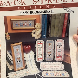 Può includere: Una pagina di un libro di artigianato intitolato "Back Street Designs Basic Bookmarks II" con disegni di segnalibri a punto croce. I disegni includono pezzi incorniciati, segnalibri con mele, mucche e un gallo, e il testo "Once upon a time."