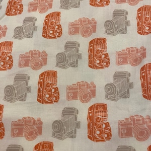 Puede incluir: Tela blanca con un patrón repetido de cámaras vintage en naranja y gris. Las cámaras son ilustraciones estilizadas, con varios diseños y detalles. La tela tiene un aspecto suave y ligeramente texturizado.