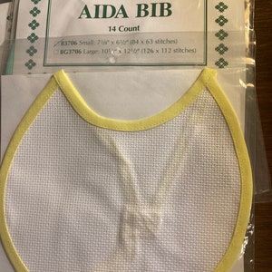May include: Aida bib for cross-stitch, with a white fabric and a yellow trim. The package includes the text "AIDA BIB" and size details: Small 7.25" x 6.5" and Large 10.5" x 12.5". The bib is packaged in clear plastic.