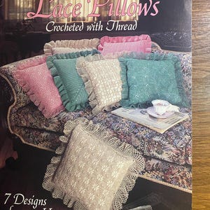 Pode incluir: Uma capa de revista com uma coleção de almofadas de renda de crochê em várias cores, incluindo rosa, verde-azulado e creme. O texto da capa diz "Lace Pillows Crocheted with Thread" e "7 Designs for your Home".
