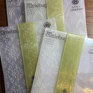 Puede incluir: Una colección de carpetas de relieve Cricut Cuttlebug con varios diseños, incluyendo patrones florales y geométricos. Las carpetas muestran el logotipo de Anna Griffin. Los colores son transparente, blanco y verde claro. Medidas no aplicables.