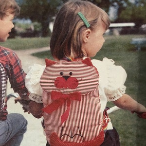 May include: A red and white striped cat-shaped backpack with a red bow and face details. The backpack is worn by a child, with a green hair clip visible. The backpack has a rainbow strap and is on a child's back.