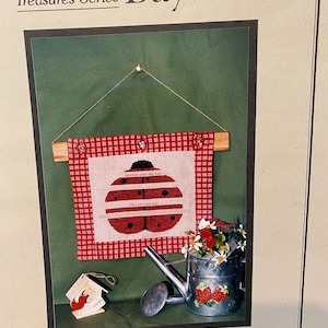 Puede incluir: Un patrón de punto de cruz con una mariquita roja con puntos negros sobre un fondo blanco. La mariquita está rodeada por un borde de cuadros rojos y blancos. El patrón está enmarcado y colgado en una pared. El texto "SUMMER SAYS" está bordado en el cuerpo de la mariquita. La imagen también incluye una pequeña casita de pájaros de madera y una regadera de metal con fresas pintadas en ella.