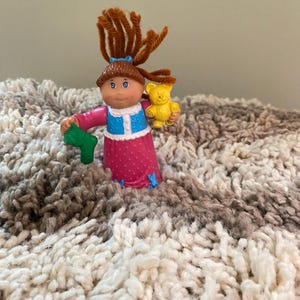 May include: A vintage Cabbage Patch Kids figurine. The doll has brown pipe cleaner hair, blue eyes, and is wearing a pink dress with white polka dots. It holds a green object in one hand and a yellow teddy bear in the other.