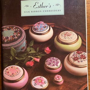 May include: A book titled "Esther's Silk Ribbon Embroidery" showcasing a selection of small, round boxes adorned with colourful silk ribbon embroidery. The boxes are displayed on a wooden tray with a floral pattern.