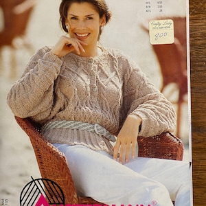 May include: A woman wearing a beige knitted sweater with a cable knit pattern, a white belt, and white trousers. She is sitting in a wicker chair. The image is from a knitting magazine with the title "Stricktrends" and the name "Austermann" printed on the bottom.