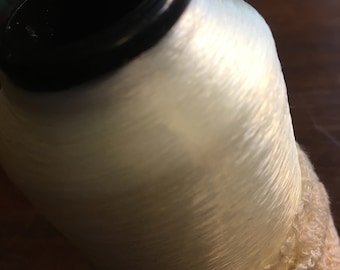 Könnte beinhalten: Eine Spule cremefarbenes Garn mit einem schwarzen Kunststoffkern. Der Faden ist eng gewickelt, und die Basis ist mit einem strukturierten, cremefarbenen Stoff umwickelt. Ideal für Näh- und Bastelprojekte.