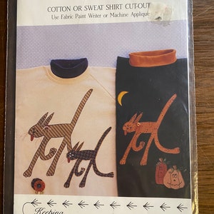 Pode incluir: Um padrão de artesanato para um recorte de camiseta de algodão ou moletom com dois gatos, abóboras e flores. O padrão é intitulado "On The Prowl" e é da coleção "Keeping You In Stitches".