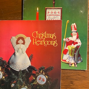 May include: Two vintage Christmas Heirloom books with cross-stitch patterns. The top book features a red candle and a small cross-stitch of a reindeer. The bottom book features a cross-stitch of an angel on top of a Christmas tree with ornaments. The text on the cover reads "Christmas Heirlooms".