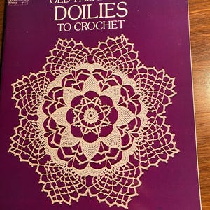 Op de afbeelding: Een boek getiteld "Old-Fashioned Doilies to Crochet" met een witte gehaakte doily op de omslag. Het boek is bewerkt door Rita Weiss en heeft een paarse omslag.