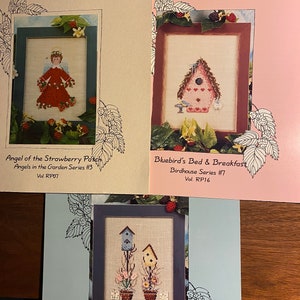 Puede incluir: Tres patrones de punto de cruz de Raspberry Patch Design Studio. El primer patrón se titula "Angel of the Strawberry Patch" y presenta un ángel con un vestido rojo. El segundo patrón se titula "Bluebird's Bed & Breakfast" y presenta una casita de pájaros con un techo azul. El tercer patrón se titula "Springtime Blooms" y presenta dos casitas de pájaros con flores.