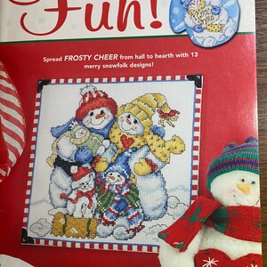 Puede incluir: Un libro de patrones de punto de cruz titulado "snow much Fun!" presenta una portada roja y blanca con un diseño de punto de cruz de muñecos de nieve. El libro incluye 13 diseños de muñecos de nieve. También se ve un adorno de guante azul con un muñeco de nieve.