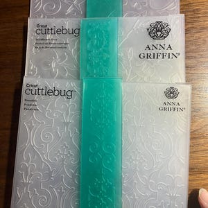 以下が含まれることがあります： Cricut Cuttlebugのエンボスフォルダーのスタック。花柄や模様のデザイン。フォルダーは透明で、エンボス加工が施されており、Anna Griffinのブランド名が記載されています。各フォルダーには、ティール色のスペーサーが付いています。