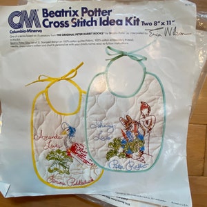 May include: A Beatrix Potter Cross Stitch Idea Kit featuring two bibs. The bibs are stamped with designs from "The Original Peter Rabbit Books." One bib features Jemima Puddleduck, and the other features Peter Rabbit. The kit includes embroidery thread and instructions.