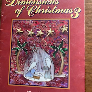Puede incluir: Un libro titulado "Dimensions of Christmas 3" con una portada roja. La portada presenta estrellas doradas, palmeras y una ilustración del nacimiento. El nombre del autor, Tony Nudson, está impreso en la parte inferior.