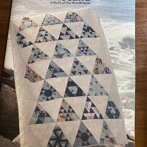 Puede incluir: Un patrón de edredón titulado "Toes in the Sand" con un diseño geométrico de triángulos en tonos azules, blancos y telas estampadas. El edredón se muestra sobre una superficie rocosa con un fondo de paisaje marino. La portada también incluye el texto "A Block of the Month Quilt" y el logotipo de Jaybird Quilts.