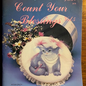 May include: A book cover featuring a white cat wearing a pink hat and a pearl necklace, sitting on a white and lace-trimmed cushion. The book title is "Count Your Blessings Vol. 5" and the author is Chris Thornton. The book cover is blue with white and blue stripes.