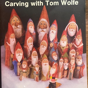 May include: A book titled Santa and His Friends: Carving with Tom Wolfe with text by Douglas Congdon-Martin. The cover features a collection of hand-carved wooden figures of Santa Claus and various companions, all wearing pointed hats and robes.