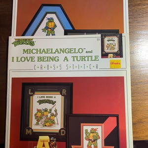 May include: Three Teenage Mutant Ninja Turtles cross-stitch kits. One kit features Michelangelo, with the text "I LOVE BEING A TURTLE". Another kit says "SLAP ME THREE, DUDE!" and includes a bonus pattern. The kits have a red and white color scheme.