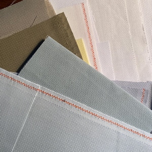 May include: A stack of different colored Aida cloth fabrics. The fabrics are arranged in a fan shape with the lightest color at the top and the darkest color at the bottom. The fabrics are all white, light gray, light brown, dark brown, and light green.