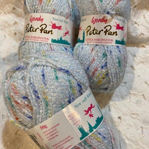 May include: Three skeins of Wendy Peter Pan Little Darlings D.K. yarn. The yarn is a light blue with colorful flecks. Each skein has a label with the brand name, a silhouette of Peter Pan, and the weight of the yarn, 50g.