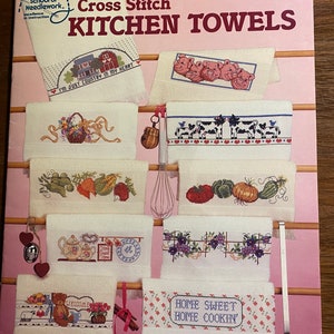 May include: A book cover with the title "Cross Stitch Kitchen Towels" by Sam Hawkins. The cover features a pink background with images of cross-stitch patterns for kitchen towels, including a farmhouse scene, pigs, cows, and a teapot.