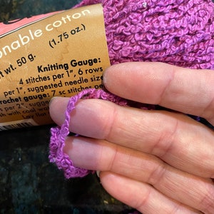 May include: A skein of purple yarn labeled "onable cotton (1.75 oz.)" with the text "wt. 50 g. Knitting Gauge: 4 stitches per 1", 6 rows per 1", suggested needle size Crochet gauge: 7 sc stitches per 2", suggesh with cool wat Lay flat erry"