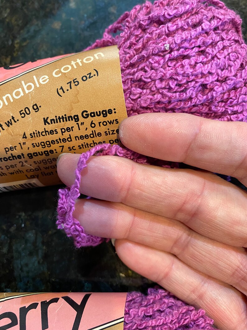 May include: A skein of purple yarn labeled "onable cotton (1.75 oz.)" with the text "wt. 50 g. Knitting Gauge: 4 stitches per 1", 6 rows per 1", suggested needle size Crochet gauge: 7 sc stitches per 2", suggesh with cool wat Lay flat erry"