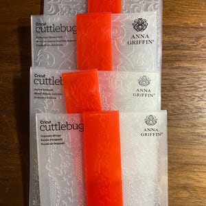 Może przedstawiać: Cztery foldery do wytłaczania Cricut Cuttlebug z kwiatowymi i geometrycznymi wzorami. Każdy folder jest przezroczysty z wzorem i trzymany razem z jasnopomarańczową opaską. Logo Anna Griffin jest widoczne na każdym folderze.