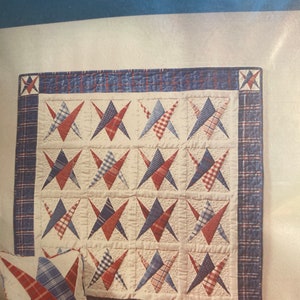 May include: A red, white, and blue patchwork quilt with a star pattern. The quilt is made of squares of different fabrics, including plaid and solid colors. The quilt is hanging on a wall.