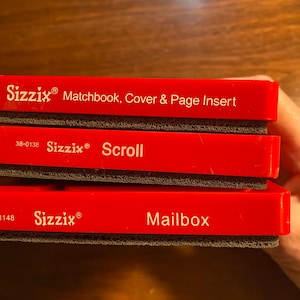 Puede incluir: Tres cajas de almacenamiento de plástico rojo con insertos de espuma negra. La caja superior dice "Sizzix Matchbook, Cover & Page Insert". La caja del medio dice "Sizzix Scroll" y la caja inferior dice "Sizzix Mailbox".