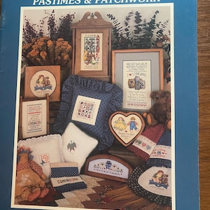 May include: A blue book cover with the text "Debra Designs PASTIMES & PATCHWORK" features various cross-stitch designs. The image showcases framed samplers, pillows, and decorative items with cross-stitch patterns, including the text "I LOVE" and "FRIENDS".