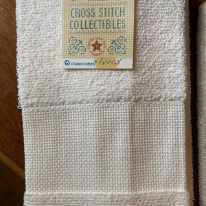 Puede incluir: Toallas de color marfil con bordado de punto de cruz y tejido texturizado. Una etiqueta dice "14 Count Border" y "Cross Stitch Collectibles". La etiqueta tambi&eacute;n presenta un dise&ntilde;o de estrella y flores, con el texto "Crafted with Pride in America."