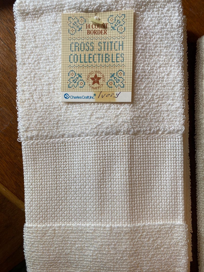 Puede incluir: Toallas de color marfil con bordado de punto de cruz y tejido texturizado. Una etiqueta dice "14 Count Border" y "Cross Stitch Collectibles". La etiqueta tambi&eacute;n presenta un dise&ntilde;o de estrella y flores, con el texto "Crafted with Pride in America."