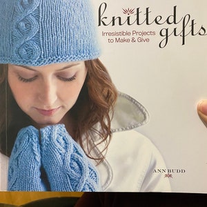 Puede incluir: Un libro titulado "Knitted Gifts: Irresistible Projects to Make & Give" de Ann Budd. La portada muestra a una mujer con un gorro y guantes de punto azules.