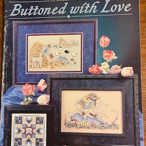 May include: A cross-stitch pattern book titled "Buttoned with Love" from the Stoney Creek Collection. The book cover features three framed cross-stitch designs: a farm scene, a girl with flowers, and a quilt block.