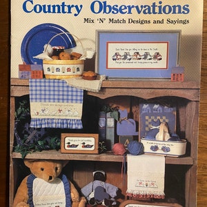 Peut inclure: Une couverture de livre avec un fond à carreaux bleus et blancs. Le titre est "Country Observations" avec le sous-titre "Mix 'N' Match Designs and Sayings". La couverture présente une variété de motifs de point de croix, notamment un ours en peluche, une poupée et un panier de fruits.