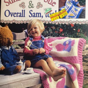 May include: A book cover titled "Sunbonnet Sue & Overall Sam" featuring a young girl and a teddy bear. The book is about crocheting afghans. The girl is sitting on a bench with a pink, white, and purple crocheted blanket.