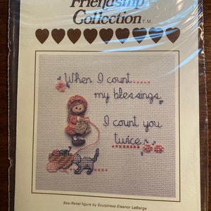Puede incluir: Patrón de punto de cruz con una niña que sostiene un gato y lana con el texto "When I count my blessings I count you twice". El patrón es parte de la colección Friendship.